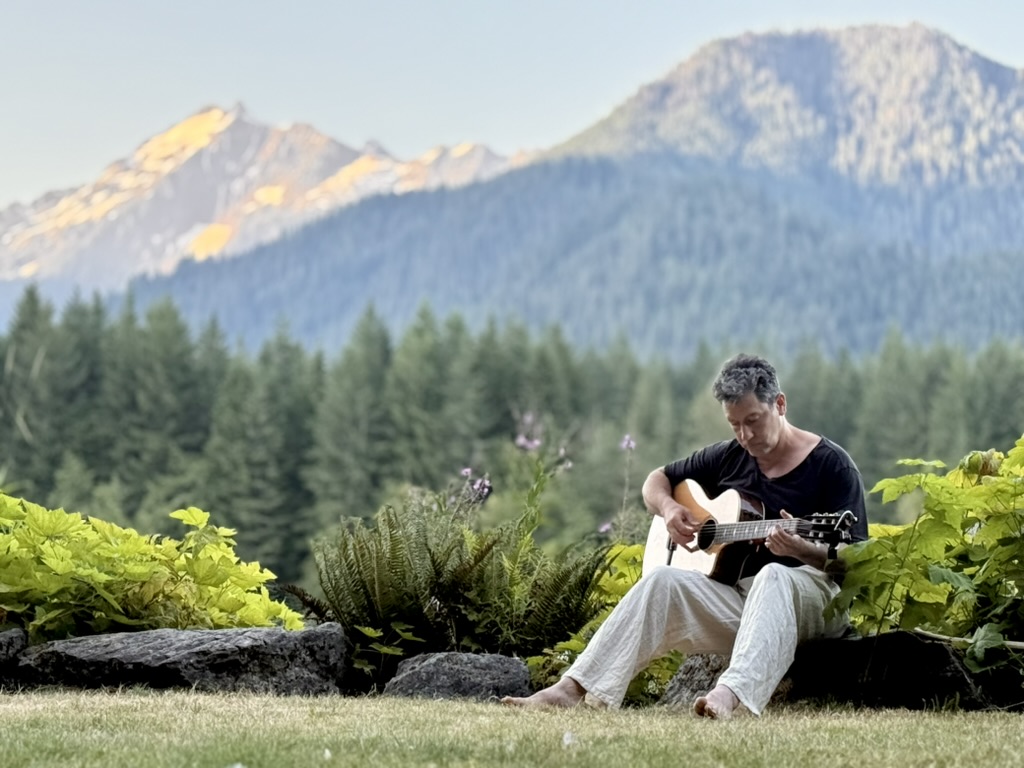 Chris Weber playing guitar barefoot, mountains behind